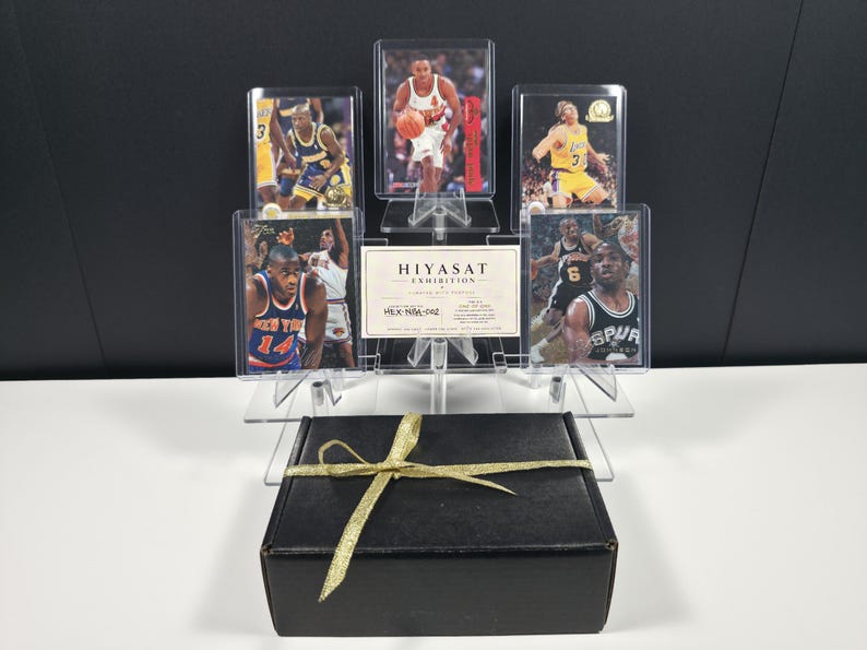 May include: A black rectangular gift box tied with a gold ribbon sits in front of a clear acrylic display stand holding five basketball trading cards and a white rectangular card with the text HIYASAT EXHIBITION and HEX-NBA-002 printed on it.