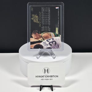 May include: A Detlef Schrempf basketball trading card numbered 130, encased in a clear plastic protective sleeve. The card back displays player statistics and team information. A white rectangular sign labeled HIYASAT EXHIBITION HEX-NBA-001 sits in front.