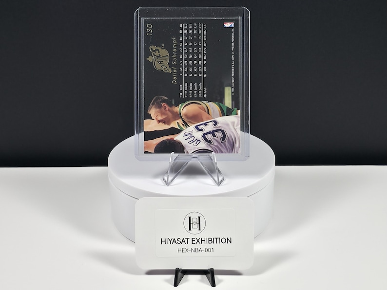 May include: A Detlef Schrempf basketball trading card numbered 130, encased in a clear plastic protective sleeve. The card back displays player statistics and team information. A white rectangular sign labeled HIYASAT EXHIBITION HEX-NBA-001 sits in front.