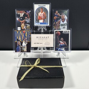Op de afbeelding: Een zwarte rechthoekige geschenkdoos met een gouden strik staat voor een doorzichtige acryl displaystandaard met vijf basketbal-verzamelkaarten en een witte kaart met de tekst HIYASAT EXHIBITION CERTIFIED WITH PASSION.