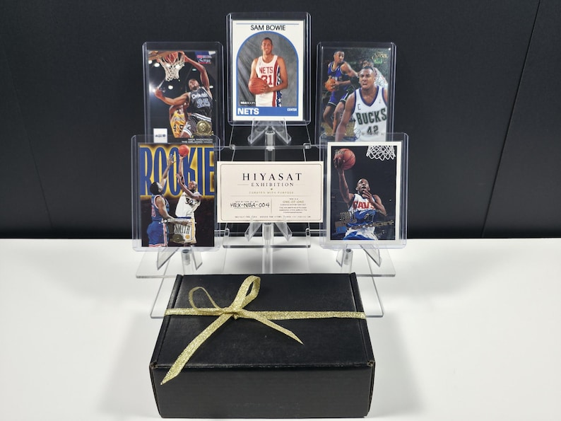 Op de afbeelding: Een zwarte rechthoekige geschenkdoos met een gouden strik staat voor een doorzichtige acryl displaystandaard met vijf basketbal-verzamelkaarten en een witte kaart met de tekst HIYASAT EXHIBITION CERTIFIED WITH PASSION.