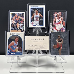 K&ouml;nnte beinhalten: Ein transparenter, gestufter Acryl-Displayst&auml;nder mit f&uuml;nf Basketball-Sammelkarten und einer zentralen rechteckigen Karte mit der Aufschrift HIYASAT EXHIBITION CURATED WITH PURPOSE HEX-NBA-003. Die Karten zeigen verschiedene Spieler in Aktion.