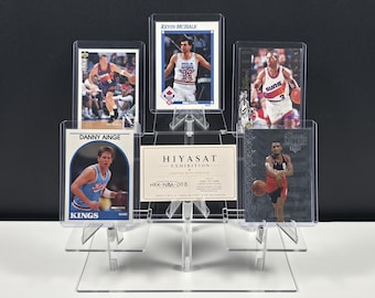 May include: A clear acrylic tiered display stand holding five basketball trading cards and a central rectangular card labeled HIYASAT EXHIBITION CURATED WITH PURPOSE HEX-NBA-003. The cards feature various players in action poses on the court.