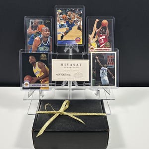 K&ouml;nnte beinhalten: Eine schwarze Geschenkbox mit goldenem Band steht unter einem transparenten Acryl-Displayst&auml;nder, der f&uuml;nf Basketball-Sammelkarten und eine rechteckige Karte mit der Aufschrift HIYASAT EXHIBITION HEX-NBA-006 enth&auml;lt. Die Karten zeigen verschiedene Spieler.