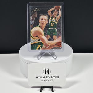 May include: A basketball trading card featuring a player in a green and yellow Sonics uniform with the number 11, encased in a clear protective sleeve. A white rectangular card in front displays the text HIYASAT EXHIBITION HEX-NBA-001.
