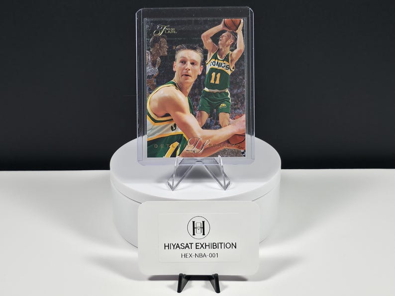 May include: A basketball trading card featuring a player in a green and yellow Sonics uniform with the number 11, encased in a clear protective sleeve. A white rectangular card in front displays the text HIYASAT EXHIBITION HEX-NBA-001.