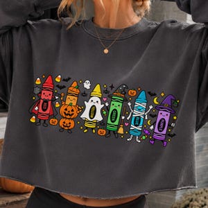 May include: A dark grey crewneck sweatshirt featuring a graphic print of six colourful crayon characters dressed in Halloween costumes, including a vampire, a pumpkin, a ghost, a witch, a mummy, and a wizard, surrounded by small bats and sweets.