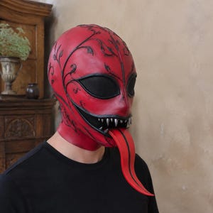 Peut inclure: Un masque intégral rouge vif orné de motifs de vignes noirs, de grands couvre-yeux en maille noire, une large bouche aux dents argentées pointues et une longue langue rouge saillante qui descend du centre de la bouche.