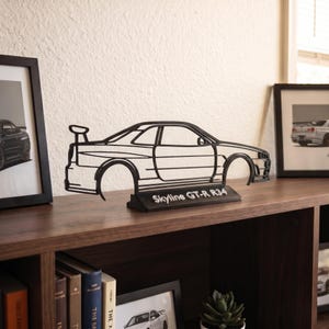 Può includere: Una scultura in filo metallico nero che raffigura la silhouette di un'auto Nissan Skyline GT-R R34 montata su una base rettangolare. La base presenta il testo bianco Skyline GT-R R34. La scultura mostra il profilo laterale con alettone.