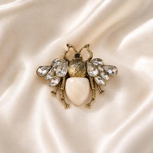 May include: A bee-shaped brooch featuring an antique gold-tone metal body with textured detailing. The insect design includes clear, faceted crystal wings and a smooth, opaque cream-coloured oval stone forming the lower abdomen section.