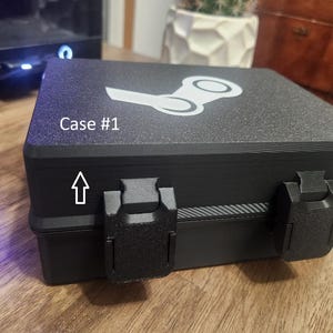 May include: A black rectangular carrying case with a textured finish, featuring two front latches and a white Steam logo printed on the lid. The text Case #1 is printed on the top left corner, accompanied by a white arrow pointing toward the latch.