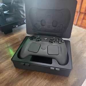 May include: A matte black video game controller with dual trackpads, analog sticks, and face buttons, resting inside a custom-fitted black hard-shell carrying case with a molded interior that matches the shape of the controller.