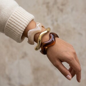 Peut inclure: Un ensemble de trois bracelets rigides ondulés portés au poignet. Le premier bracelet est en résine marbrée beige clair, le deuxième est en métal doré poli et le troisième est en résine translucide brun foncé. Chaque bracelet a une forme ondulée.