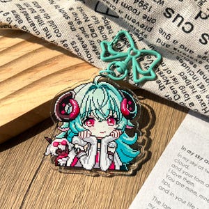 Peut inclure: Un porte-clés en acrylique transparent représentant un personnage en pixel art aux cheveux bleu sarcelle, aux yeux rouges et aux cornes sombres. Le personnage porte un manteau blanc et est accompagné d'une petite créature blanche avec un fermoir nœud.