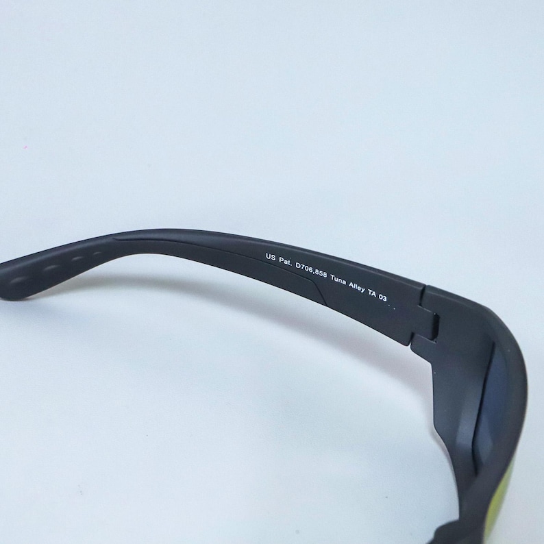 Puede incluir: Un par de gafas de sol negro mate con un dise&ntilde;o de montura curva. El interior de la patilla tiene impreso el texto US Pat. D706,858 Tuna Alley TA 03, que indica el modelo espec&iacute;fico y la informaci&oacute;n de patente de esta montura.