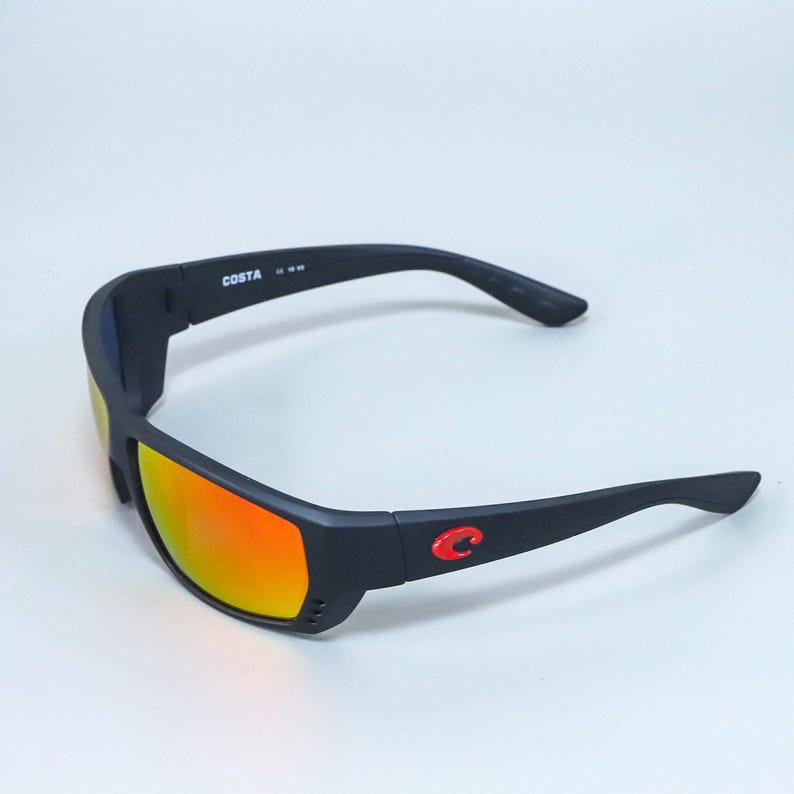 Puede incluir: Gafas de sol negras con lentes de espejo naranja y montura negra mate. La patilla presenta el texto blanco COSTA y un emblema de logotipo rojo. La montura incluye peque&ntilde;as ranuras de ventilaci&oacute;n cerca de las lentes en las esquinas.