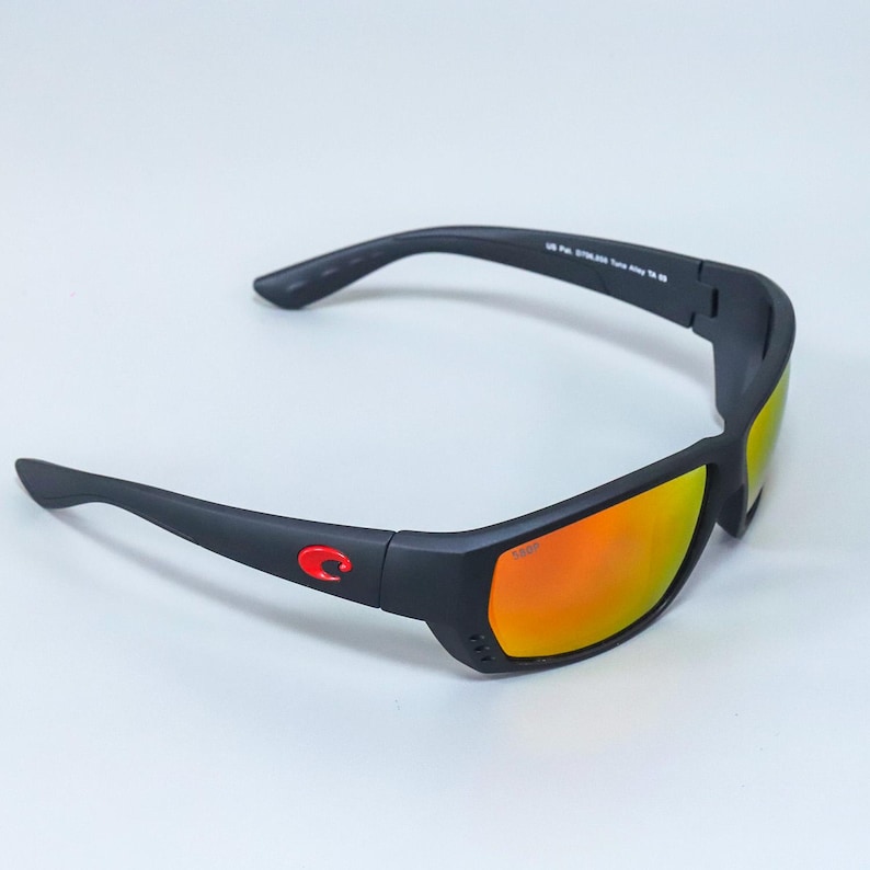 Puede incluir: Gafas de sol negro mate con lentes naranjas reflectantes y un logotipo rojo en la patilla. La montura incluye peque&ntilde;as ranuras de ventilaci&oacute;n cerca de la lente y el texto US Pat. D706,855 Tuna Alley TA 09 en la patilla interior.