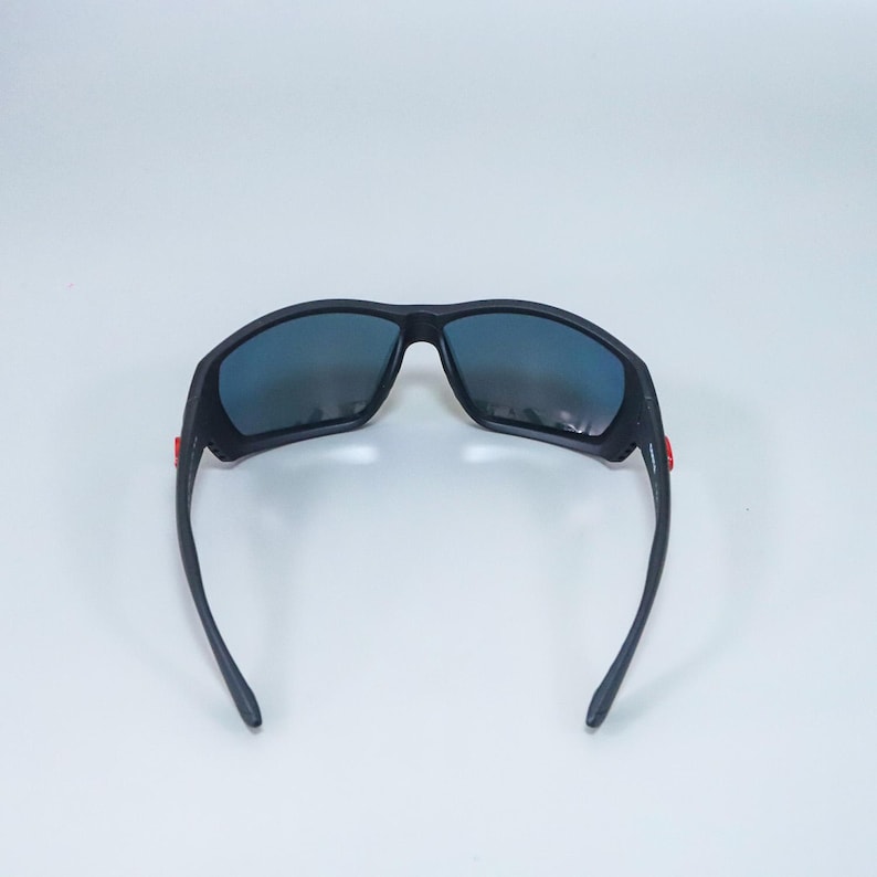 Puede incluir: Gafas de sol negras con montura de acabado mate y lentes oscuras y reflectantes. Las patillas presentan peque&ntilde;os detalles rectangulares de color rojo brillante cerca de las bisagras, lo que aporta un sutil contraste al dise&ntilde;o oscuro de las gafas.