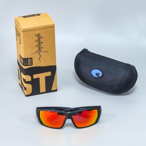 Puede incluir: Gafas de sol negras con lentes naranjas reflectantes, un estuche r&iacute;gido negro con cremallera y un emblema azul, y una caja de cart&oacute;n rectangular con el texto Costa, Born on the Water y una ilustraci&oacute;n gr&aacute;fica de un anzuelo de pesca.