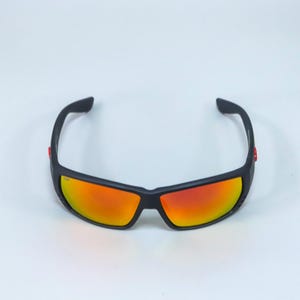 Puede incluir: Gafas de sol deportivas con montura negra mate y lentes reflectantes de color naranja rojizo. El dise&ntilde;o envolvente incluye peque&ntilde;os detalles rojos en las patillas exteriores, ofreciendo un aspecto elegante con un amplio campo de visi&oacute;n.