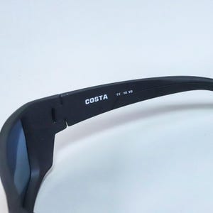 Puede incluir: Gafas de sol negras con acabado mate que presentan el nombre de la marca COSTA impreso en blanco en la patilla interior. La patilla tambi&eacute;n muestra el texto CE 18 V3. La montura tiene un dise&ntilde;o curvo con una bisagra cerca de la sien.