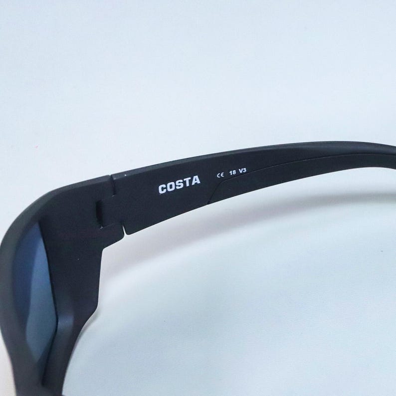 Puede incluir: Gafas de sol negras con acabado mate que presentan el nombre de la marca COSTA impreso en blanco en la patilla interior. La patilla tambi&eacute;n muestra el texto CE 18 V3. La montura tiene un dise&ntilde;o curvo con una bisagra cerca de la sien.
