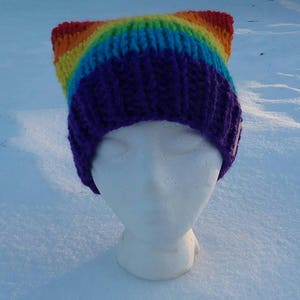 May include: A rainbow knit beanie with cat ears. The beanie is purple, blue, green, yellow, orange, and red. The beanie is on a white mannequin head.