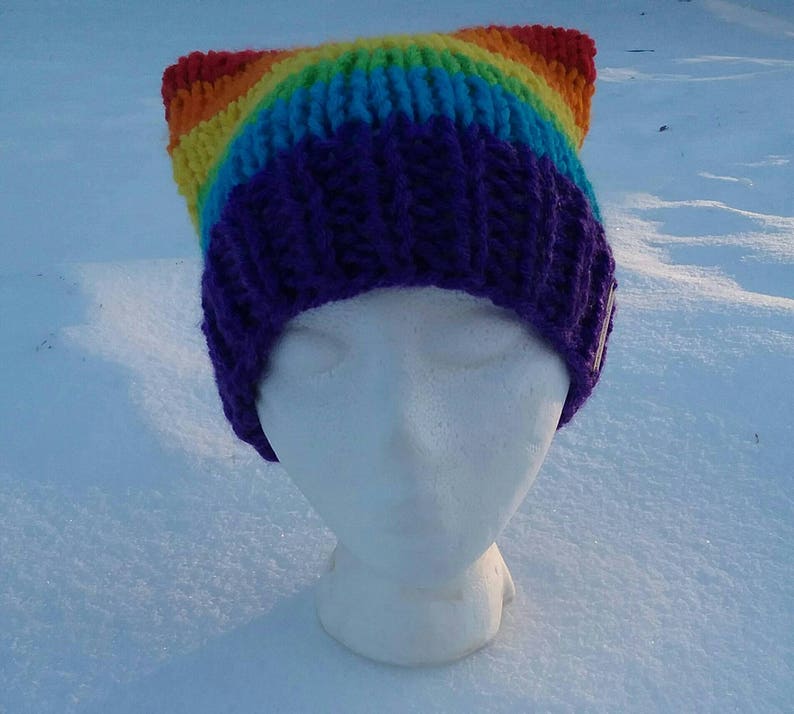 May include: A rainbow knit beanie with cat ears. The beanie is purple, blue, green, yellow, orange, and red. The beanie is on a white mannequin head.