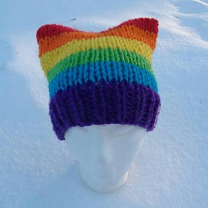 May include: A rainbow knit hat with a purple brim. The hat is on a white styrofoam head and is sitting on a snowy surface.