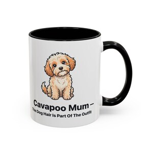 May include: A white ceramic mug with a black interior and handle. The front features a cartoon illustration of a tan and white Cavapoo puppy. Below the puppy, the text reads: Cavapoo Mum - The Dog Hair Is Part Of The Outfit in black font.