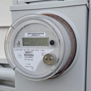May include: A gray digital electricity meter with a clear circular cover, mounted on a gray rectangular housing. The device features a digital display, a barcode, and text including CL200 240V 3W TYPE CS2M 30TA 1.0Kh, DUKE ENERGY, and OpenWay.
