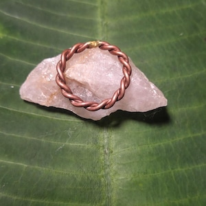 May include: A circular ring crafted from twisted copper wire, featuring a small gold-colored accent at the top. The ring is positioned resting on a translucent, pale pink piece of raw quartz crystal, showcasing a metallic and earthy aesthetic.