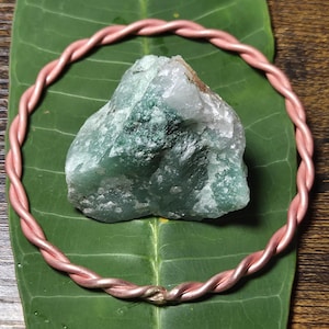 May include: A circular bracelet made of twisted copper wire encircling a rough, jagged piece of green and white aventurine stone. The stone features a mix of translucent white quartz and opaque green mineral deposits with a natural, unpolished texture.
