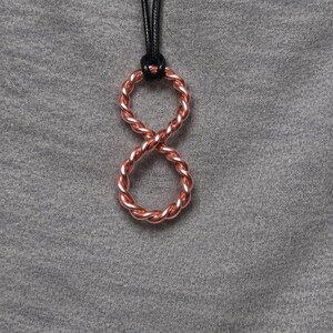 May include: A copper-colored pendant shaped like an infinity symbol, constructed from two twisted wire loops joined together. The pendant hangs from a thin, smooth black cord, creating a continuous loop design that rests against a gray fabric surface.