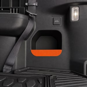 May include: The interior cargo area of a vehicle features a black plastic side panel with a recessed storage cubby highlighted by a bright orange base. An integrated light, a power outlet, a seatbelt strap, and a floor mat are visible.