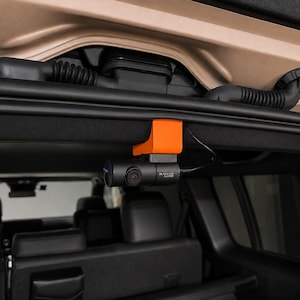 Peut inclure: Une caméra de tableau de bord cylindrique noire BlackVue Full HD fixée au toit intérieur d'un véhicule à l'aide d'un support en plastique orange vif. La caméra présente un petit objectif à l'avant et un fin câble d'alimentation noir sur le côté.