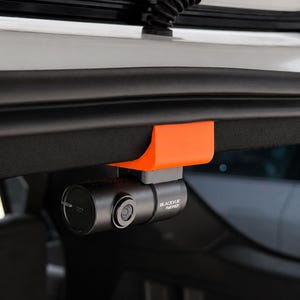 May include: A cylindrical black BlackVue Full HD dash camera mounted to the interior trim of a vehicle using a bright orange plastic bracket. The camera features a circular lens on the side and a circular end cap with a small white logo.
