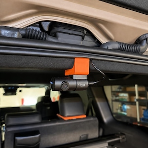 May include: A cylindrical black BlackVue Full HD dash camera mounted to the interior roof lining of a vehicle using a bright orange plastic bracket. A thin black power cable extends from the side of the camera, which features a small blue light.