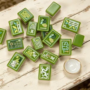 May include: A collection of rectangular green Mahjong tiles featuring various illustrations including a dragon, peacock, bamboo, and the text JOKER, NORTH, and EAST, alongside a small white ceramic bowl with a gold rim, scattered on a white surface.