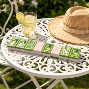 May include: A row of eight rectangular mahjong tiles arranged on a white table. The tiles feature green and pink color blocks with printed designs including birds, the word EAST with an arrow, a dragon, and the word JOKER with a hand illustration.