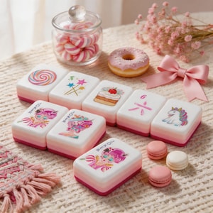 May include: A set of white mahjong tiles with pink gradient bases, featuring colorful illustrations of lollipops, stars, cakes, unicorns, and stylized characters. The tiles are arranged alongside a glass jar of pink candies, a donut, and a pink bow.