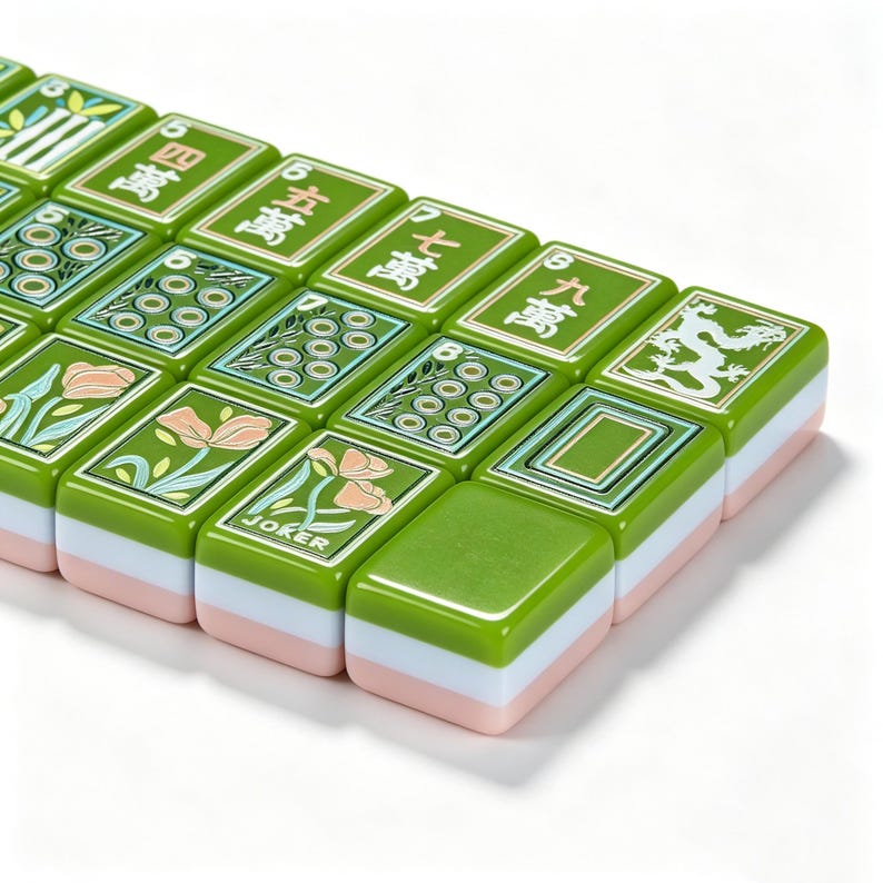 May include: A collection of rectangular Mahjong tiles featuring a green top surface with various engraved designs, including Chinese characters, floral patterns, a dragon, and the word JOKER. Each tile has a layered construction with green, white, and pink sides.