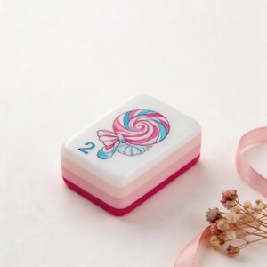 May include: A rectangular white tile with a layered pink and magenta base. The top surface features a graphic illustration of a pink and blue swirled lollipop and the number 2 printed in blue on the left side of the tile surface.