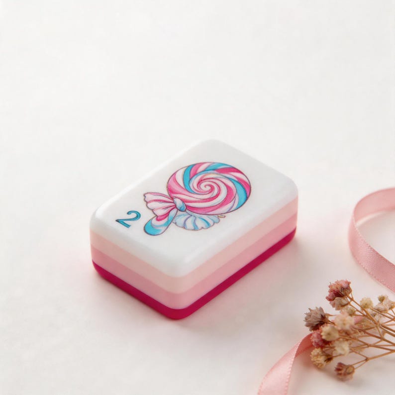 May include: A rectangular white tile with a layered pink and magenta base. The top surface features a graphic illustration of a pink and blue swirled lollipop and the number 2 printed in blue on the left side of the tile surface.