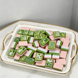 May include: A collection of rectangular mahjong tiles featuring a pastel pink and green color scheme, displayed on a white rectangular serving tray with gold-toned handles. The tiles include various engraved designs such as bamboo, dragons, and text.