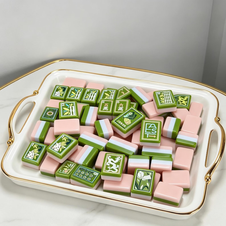 May include: A collection of rectangular mahjong tiles featuring a pastel pink and green color scheme, displayed on a white rectangular serving tray with gold-toned handles. The tiles include various engraved designs such as bamboo, dragons, and text.