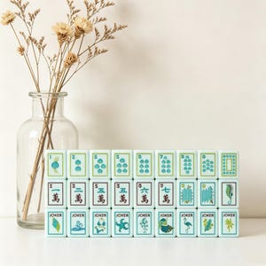 以下が含まれることがあります： 3列に並べられた水色の麻雀牌のセット。海生生物、植物、漢字の緑と青のイラストが描かれており、横には乾燥したベージュの花が入った透明なガラスの花瓶が置かれています。