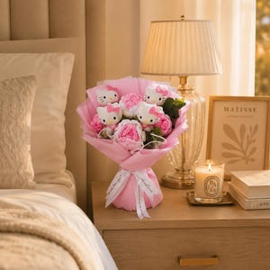 May include: A bouquet wrapped in pink paper featuring three white Hello Kitty plush figures with pink bows, interspersed with pink and white fabric flowers and green leaves. The bouquet is tied with a white ribbon featuring black text.