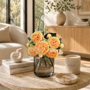 May include: A bouquet of artificial peach-colored peony flowers with green leaves arranged in a translucent dark gray glass vase. The vase sits on a round beige stone table alongside a white candle labeled JAIPUR and a small round stone tray.