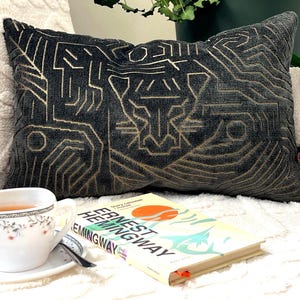 May include: A rectangular dark gray throw pillow featuring a gold embroidered geometric tiger face design. The pillow has a small black tag with a red logo on the right side. A white teacup and a book titled Ernest Hemingway are positioned in front.